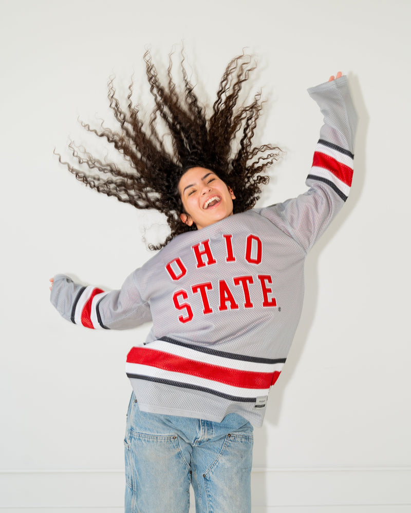 A woman with voluminous curly hair lies on her back against a white wall, wearing a gray Ohio State varsity-style sweatshirt with red and white lettering and blue jeans.
