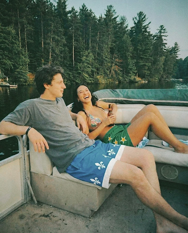 A man and a woman laughing together in a pontoon boat on a lake. The man wears a gray t-shirt, the Fleur swim trunks with white fleur-de-lis, and the girl is wearing The Miracle Man swim trunks.