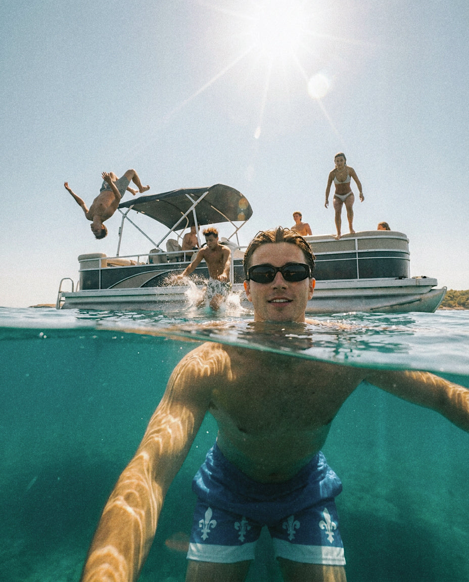 A man wearing the Fleur swim trunks and black sunglasses, swimming in clear turquoise water, with a sunny boat scene and people jumping off the pontoon behind him.