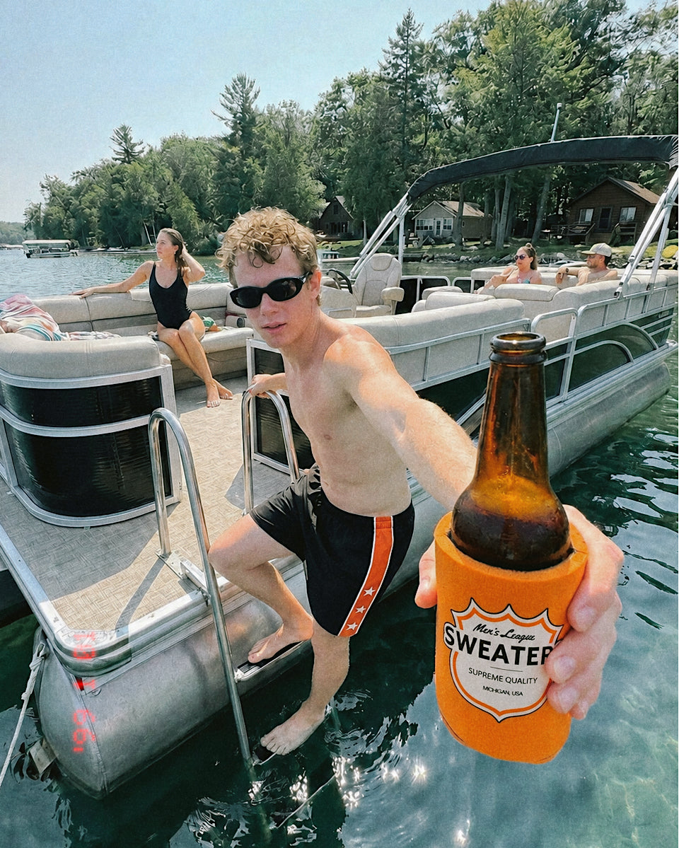 A man on a pontoon boat holds up an orange "Men's League Sweaters" beer koozie with a brown bottle inside, with other people and a lake background visible.