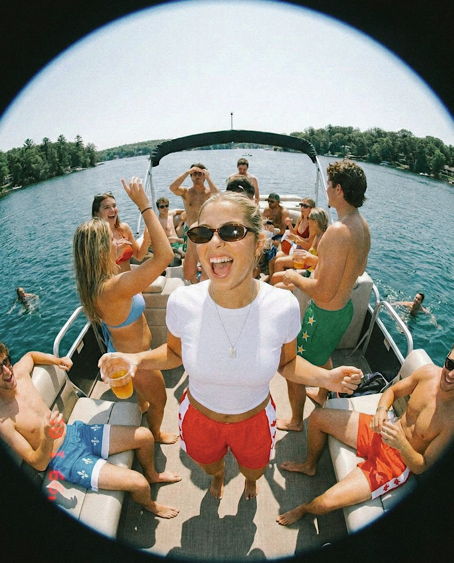 A group of friends partying on a boat with blue water and trees in the background, captured through a circular fisheye lens. A woman in the center wears a white crop top and the Maple swim trunks that are red with a white strip and three red maple leaves