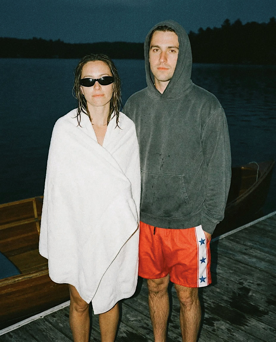 A man and a woman standing on a wooden dock at dusk by the water. The guy is wearing The Right Players swim trunks that are red with a white strip on the side with three blue stars. The woman is wrapped in a white towel and wearing black sunglasses.