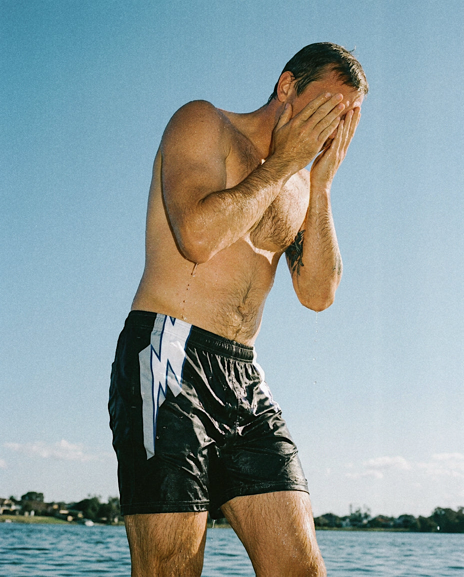 A shirtless man standing outdoors by the water wearing the Bay Bay swim trunks that are black with a white and grey lightning bolt