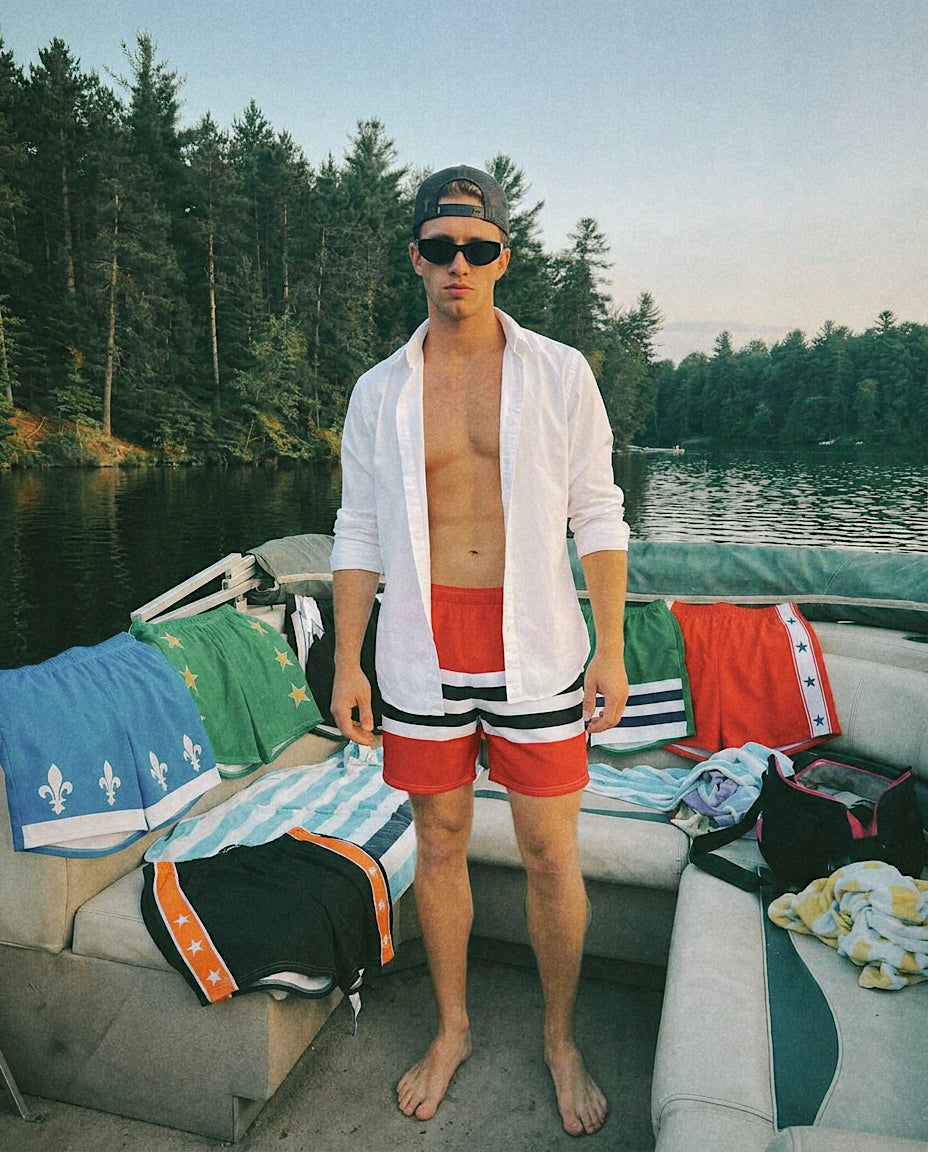 A man stands on a boat wearing The JR swim trunks that are red with black and white stripes, an open white shirt, and sunglasses. The boat has the other swim trunks and a cooler, with a forest and lake in the background.