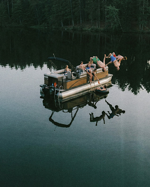 A high-angle view of a pontoon boat on a calm lake, with several people lounging and jumping off the side into the water. The background shows a dark, dense forest reflected in the water.