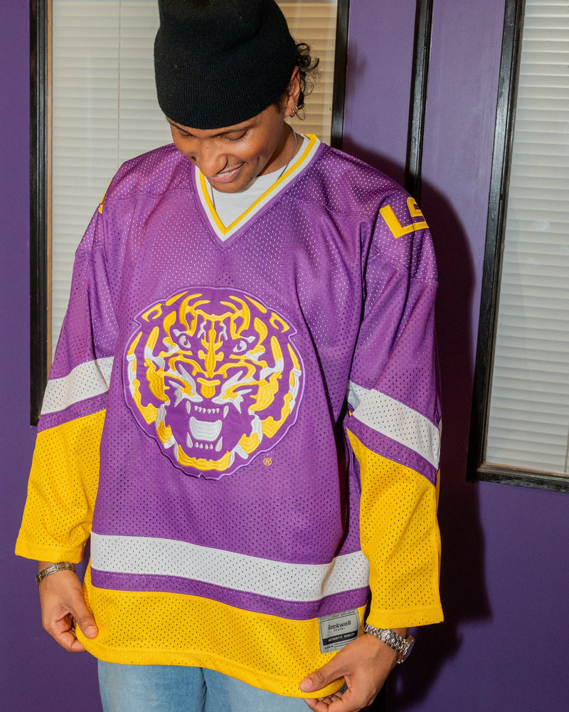 A person wearing a purple and yellow LSU Tigers hockey jersey, blue jeans, and a black beanie, standing against a purple wall with window blinds.