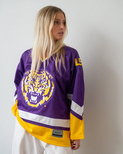 A woman wearing a purple and yellow LSU Tigers hockey jersey with white pants, standing against a plain light gray wall.