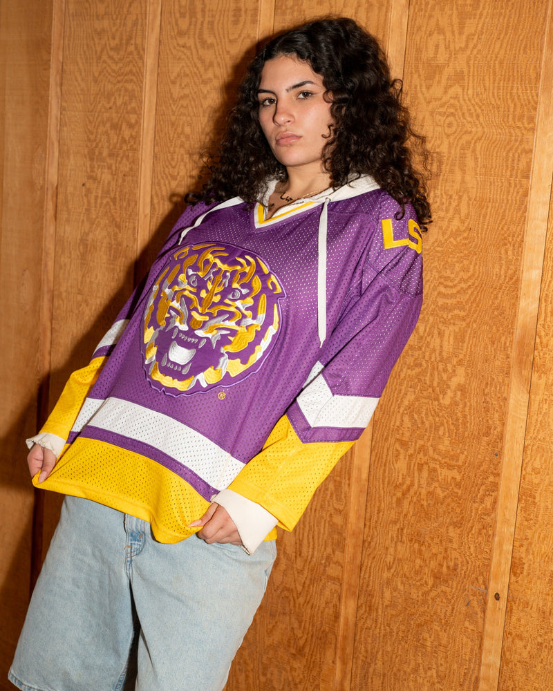 A woman wearing a purple and yellow LSU Tigers hockey jersey with a tiger graphic on the front, paired with light blue denim shorts, posing against a wood-paneled wall.