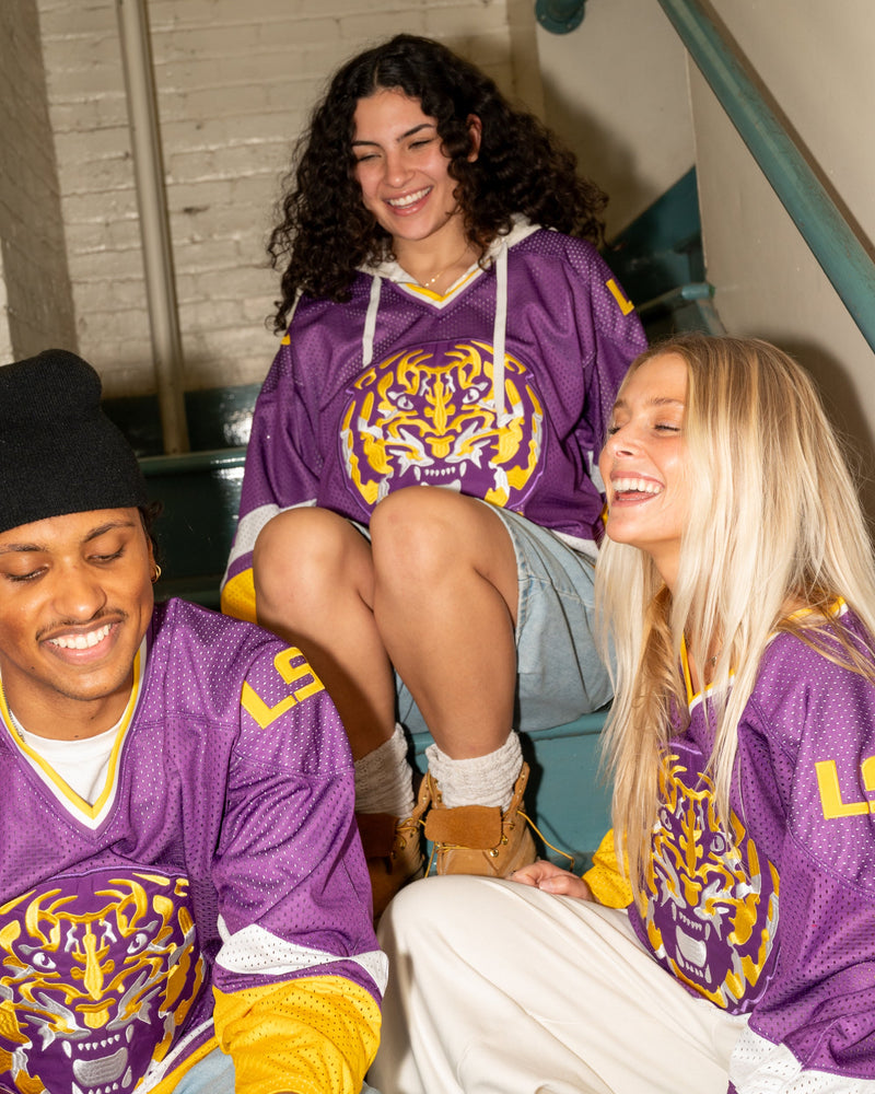 Three people wearing matching purple and yellow LSU Tigers jerseys, laughing on an indoor staircase. The jerseys feature a large tiger graphic on the front with 'LSU' lettering.
