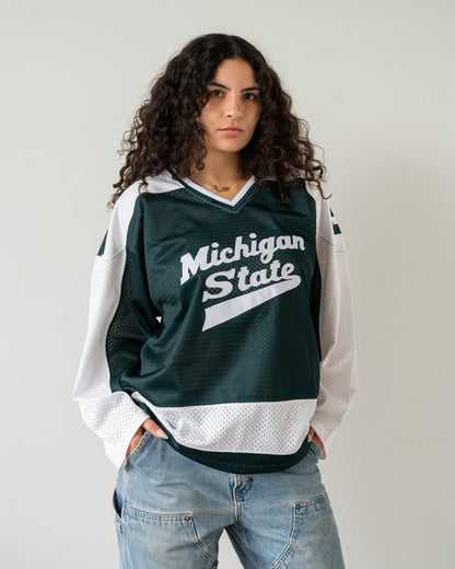 A woman wearing a dark green and white Michigan State hockey jersey with light blue jeans, standing against a plain light gray wall.