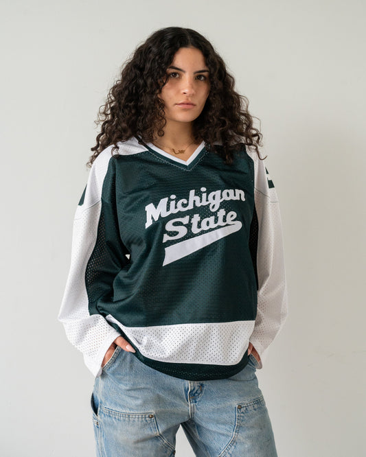 A woman wearing a dark green and white Michigan State hockey jersey with light blue jeans, standing against a plain light gray wall.