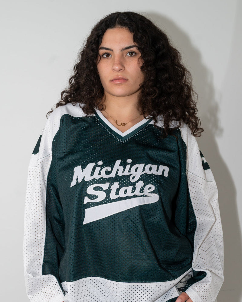 A woman with curly dark hair wearing a dark green and white Michigan State hockey jersey and light blue jeans, standing against a plain light gray wall.