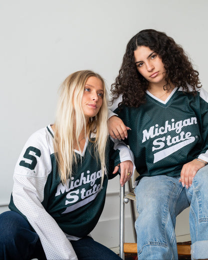 Two women wearing Michigan State hockey jerseys, one in a white and dark green long-sleeve jersey and the other in a dark green short-sleeve jersey, sitting against a plain light gray wall.