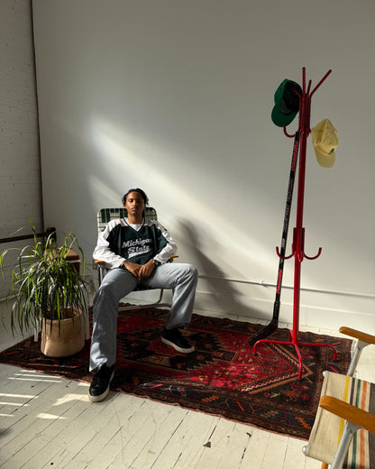 A person sits in a chair on a patterned rug in a sunlit room with a white wall, wooden floor, potted plant, and a red coat rack holding hats and a ski boot.
