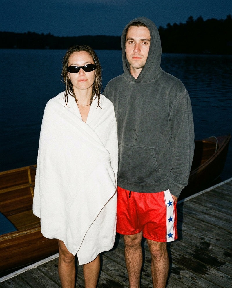 A man and a woman standing on a wooden dock at dusk by the water. The guy is wearing The Right Players swim trunks that are red with a white strip on the side with three blue stars. The woman is wrapped in a white towel and wearing black sunglasses.