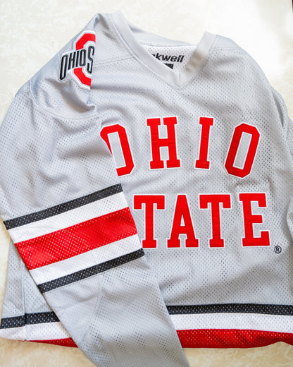 A light gray Ohio State hockey jersey laid flat on a beige upholstered surface. The jersey features 'OHIO STATE' in red block letters across the chest, with black, white, and red striped detailing on the sleeves and lower hem.