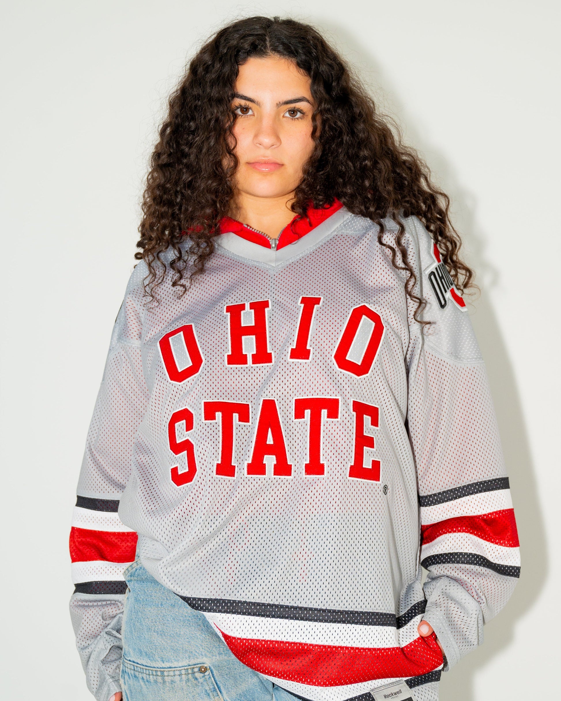 A woman with curly dark hair wearing a gray Ohio State hockey jersey with red and black accents, paired with light-wash blue jeans, standing against a plain off-white wall.
