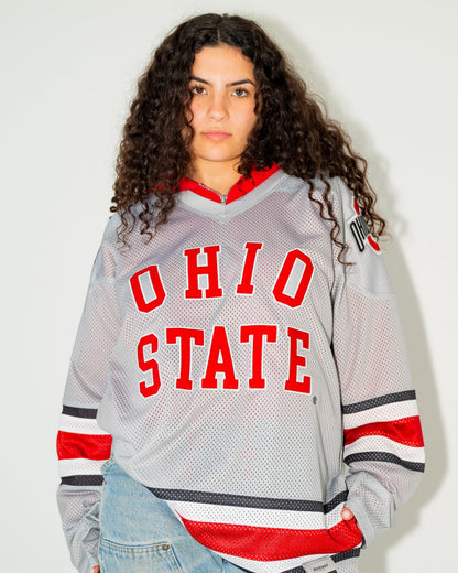 A woman with curly dark hair wearing a gray Ohio State hockey jersey with red and black accents, paired with light-wash blue jeans, standing against a plain off-white wall.