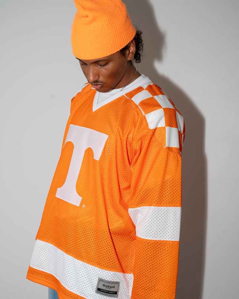 A person wearing an oversized bright orange Tennessee Volunteers mesh hockey-style jersey with white stripes and a large white 'T' logo, paired with an orange beanie and light blue jeans, standing against a plain light gray wall.