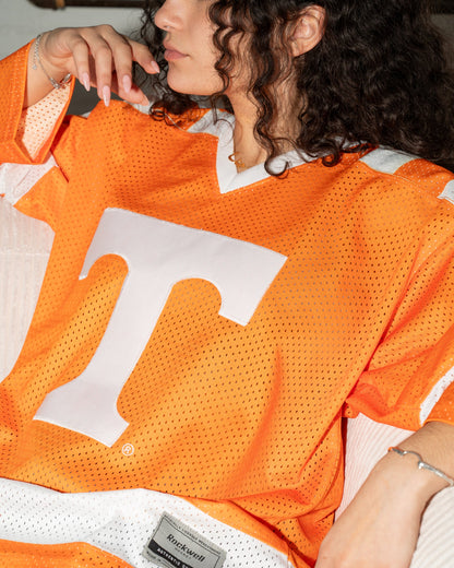 A close-up lifestyle shot of a woman wearing an orange Tennessee Volunteers mesh football jersey with a large white 'T' logo on the front. The jersey features white mesh panels and a visible 'Miz Mooz' brand patch near the hem.