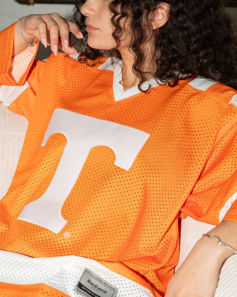 A close-up lifestyle shot of a woman wearing an orange Tennessee Volunteers mesh football jersey with a large white 'T' logo on the front. The jersey features white mesh panels and a visible 'Miz Mooz' brand patch near the hem.