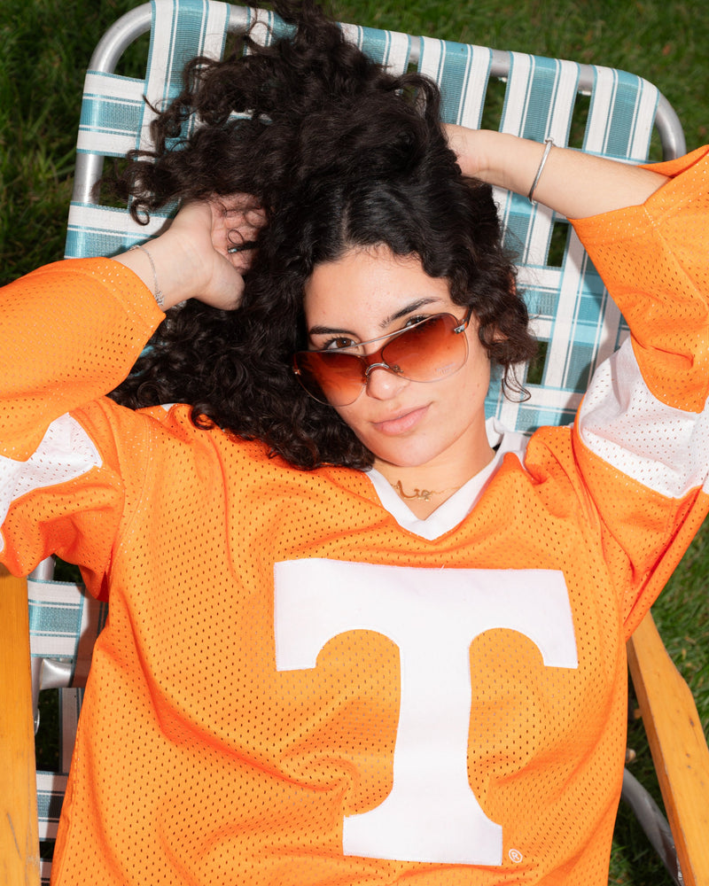 A woman wearing a bright orange Tennessee Volunteers mesh jersey with a large white 'T' logo, reclining on a green-and-white striped lawn chair outdoors.