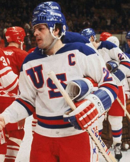 A hockey player in a white USA jersey with blue and red trim, wearing a blue helmet and gloves, shaking hands with an opponent in a red jersey on an ice rink. Other players are visible in the background.
