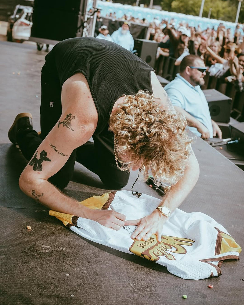 Yung Gravy kneels on a stage, signing a white sports jersey with a gold graphic. The background shows a live concert crowd and stage equipment.